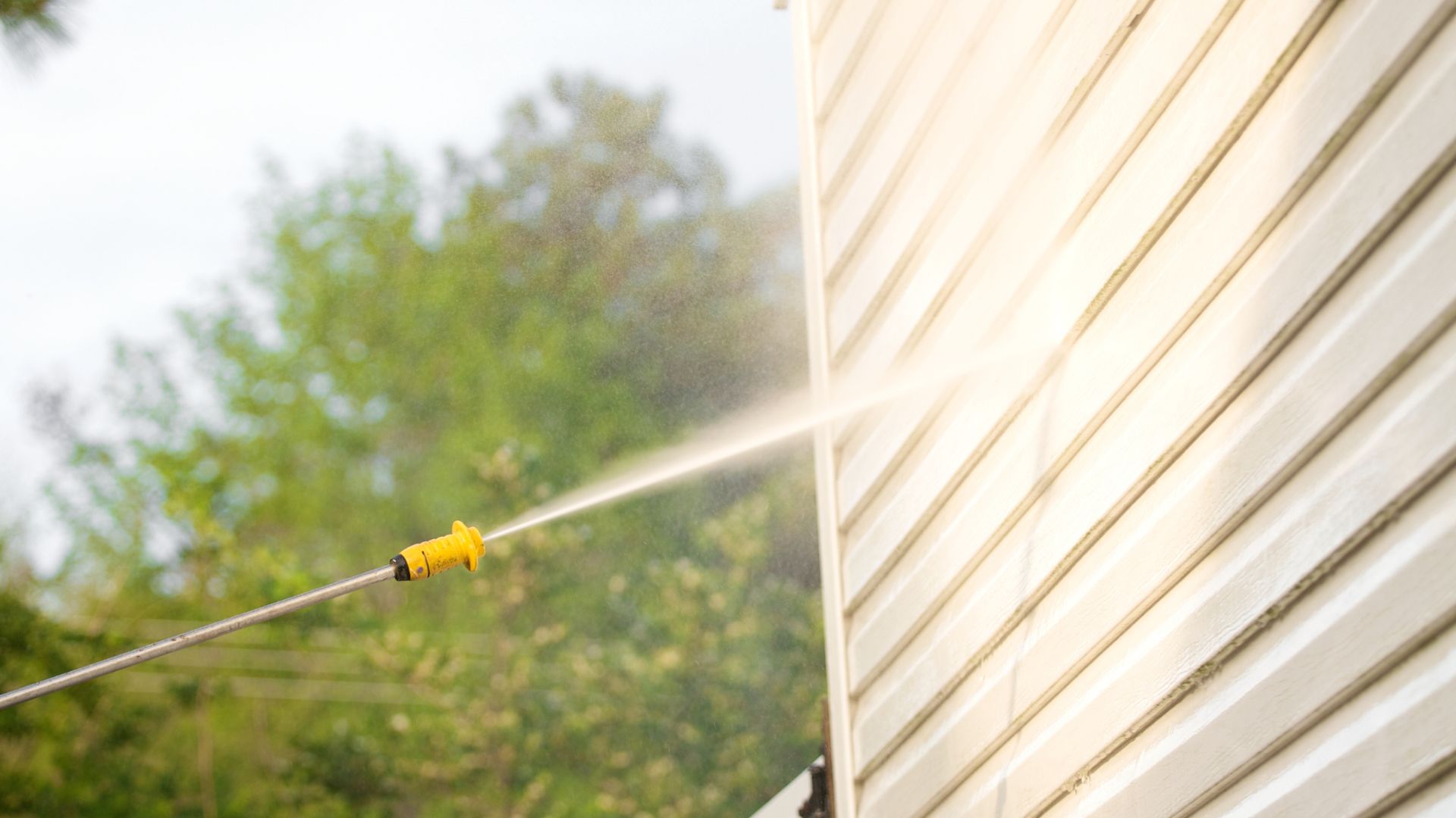 Yellow nozzle spraying water on house siding with trees in background